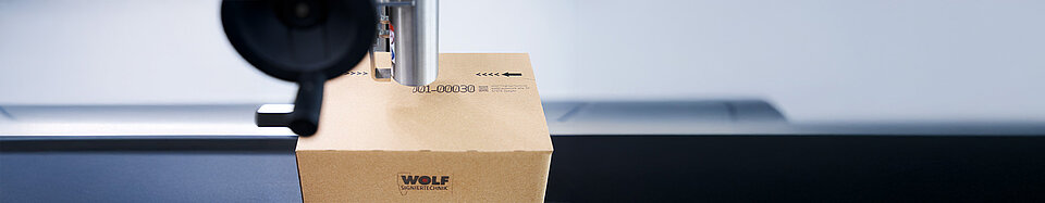 Ein Druckkopf bedruckt einen Versandkarton mit einem Barcode und dem Logo von WOLF Signiertechnik